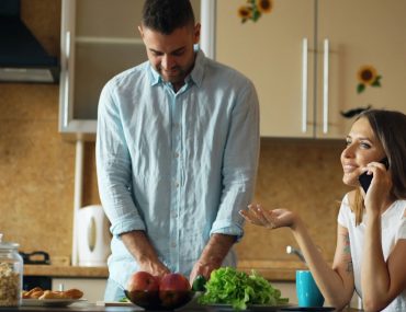 Attractive couple in the kitchen early morning. Beautiful girl chatting phone while her boyfriend cooking breakfast at home