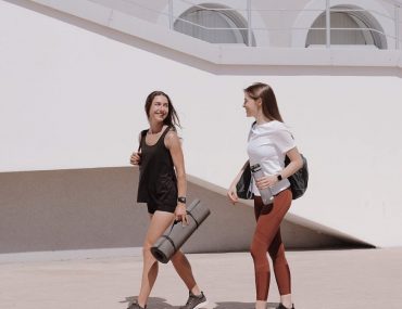 Two women in active wear walking outdoors while smiling and carrying yoga mats.