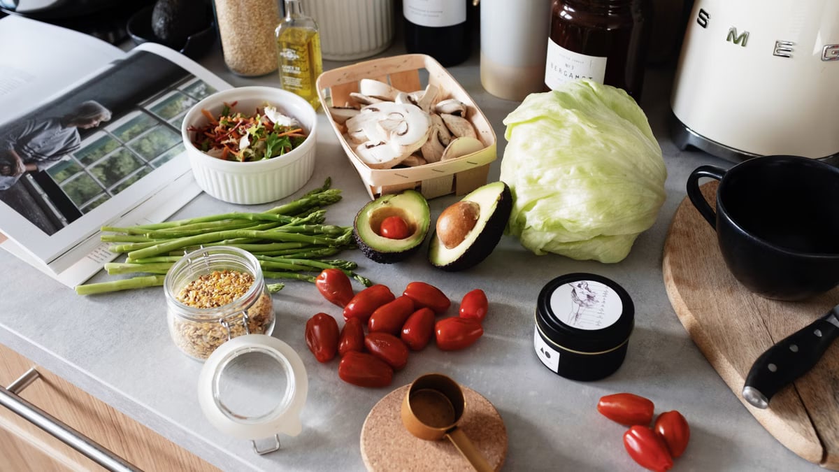 From above of various organic vegetables chopped ingredients near cutting board kitchenware appliances and trendy magazine placed on gray countertop in kitchen at home